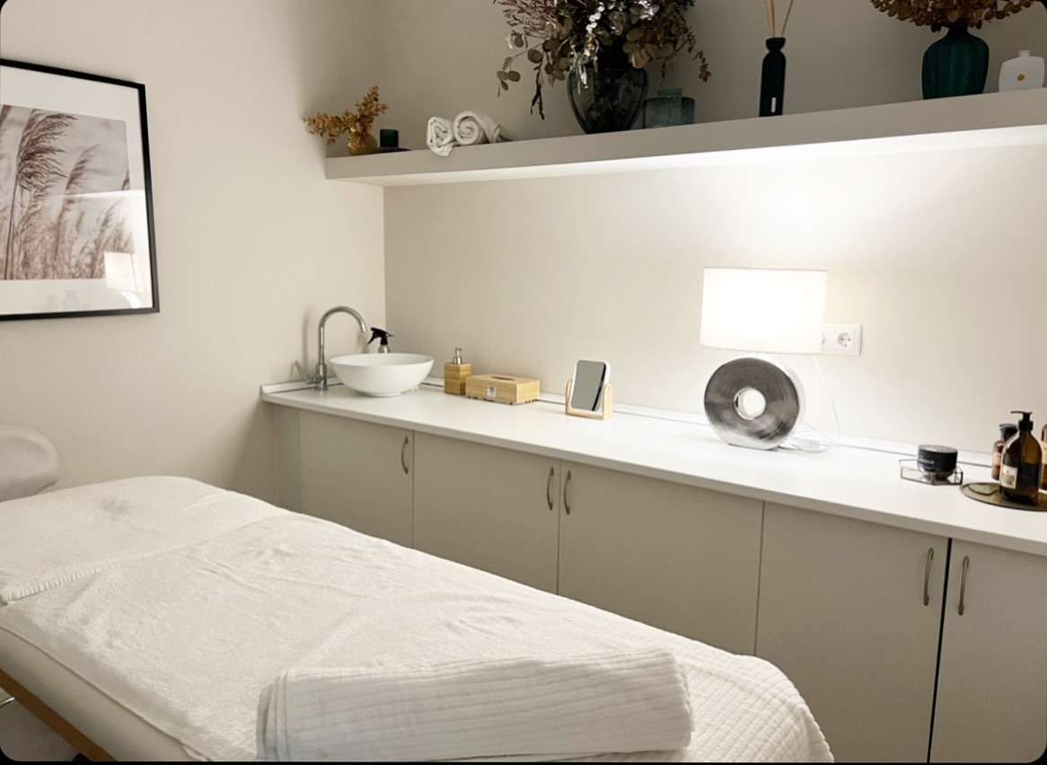 Sala de tratamientos de spa con camilla de masaje, lavabo y estante decorativo.