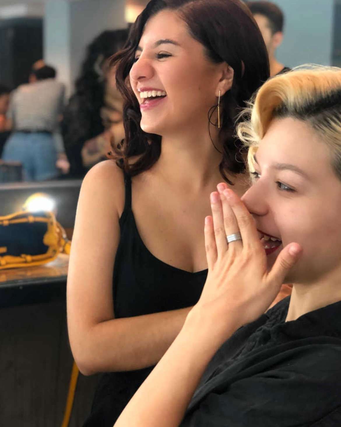 Dos personas riendo; una de ellas, con el pelo corto y rubio, se tapa la boca con la mano.