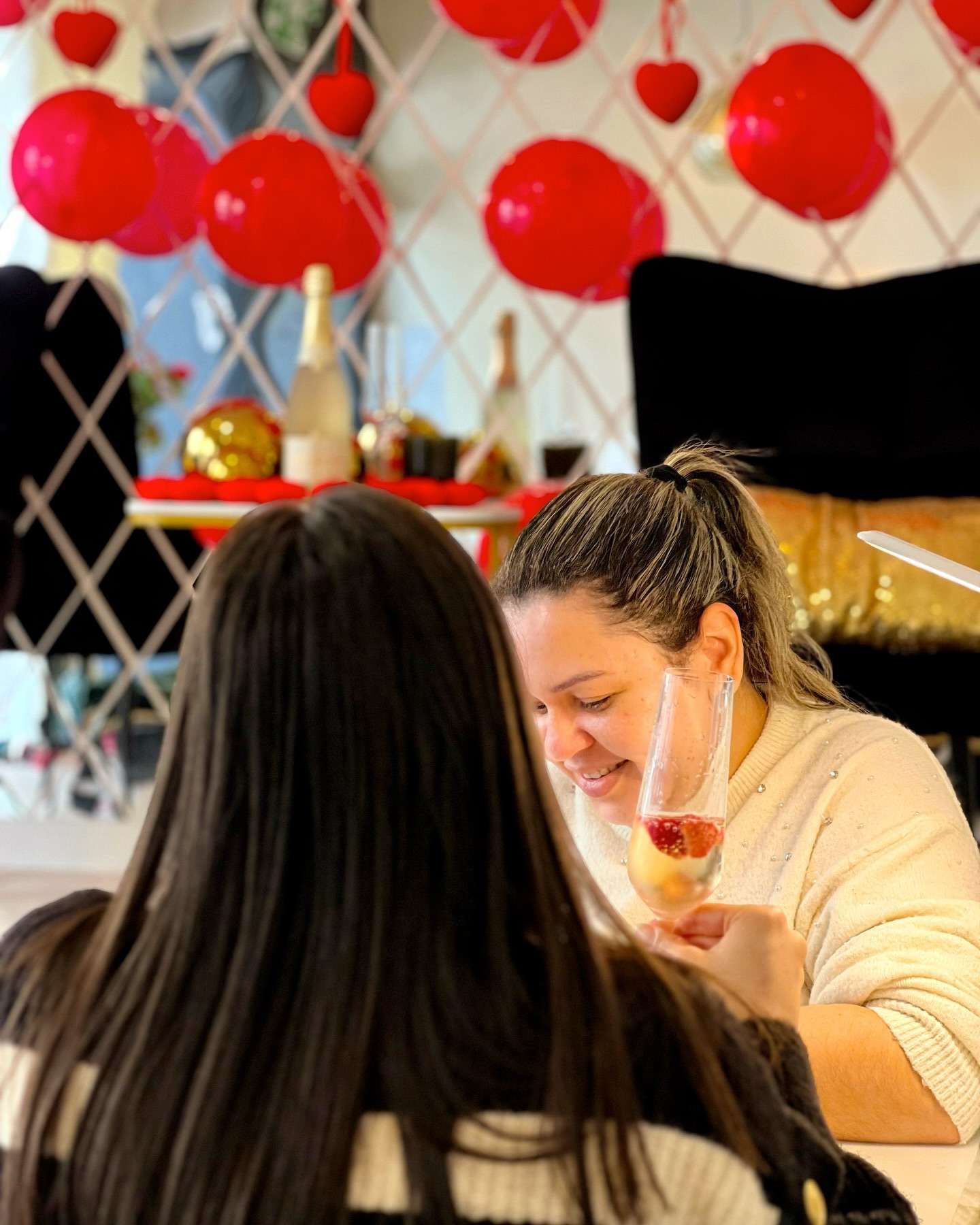 Dos mujeres sentadas a una mesa, una de ellas bebiendo una bebida rosa, decorada con globos rojos y corazones.