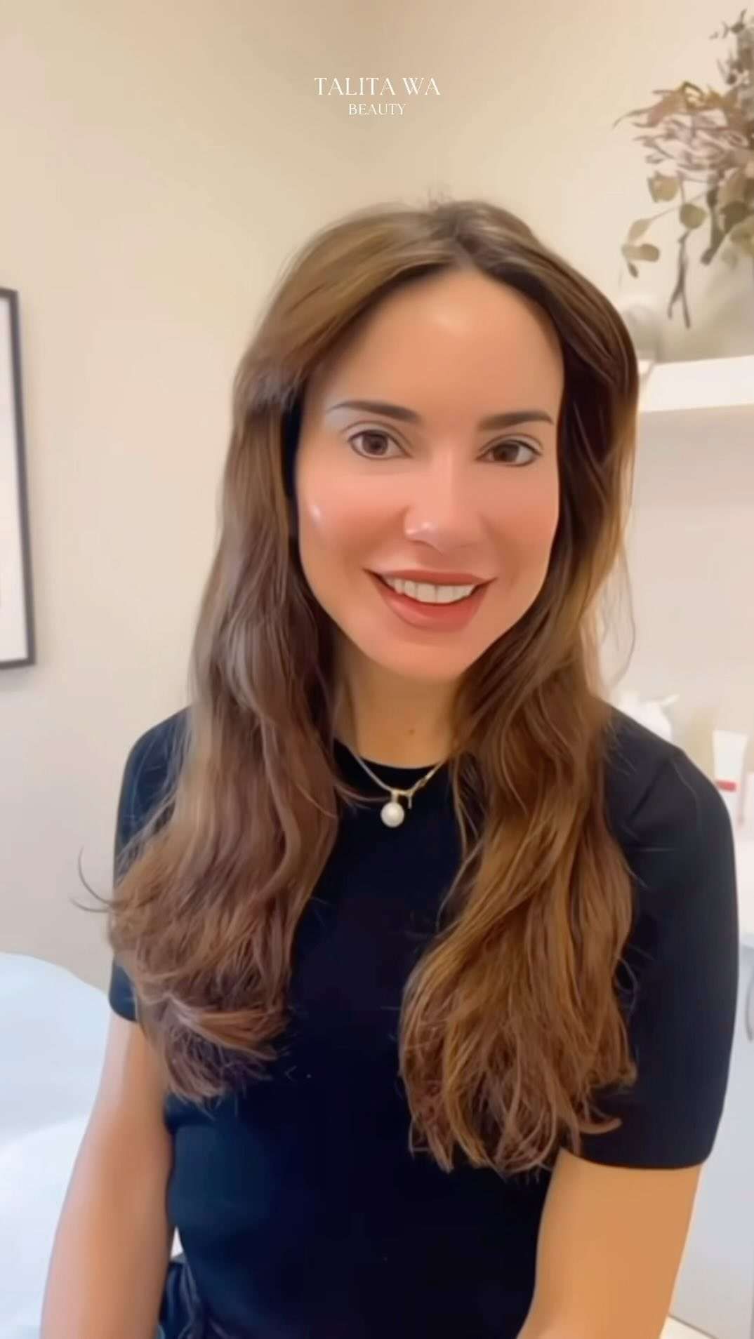 Mujer con cabello largo y castaño, sonriente, vestida con una camisa negra y un collar de perlas en una clínica.