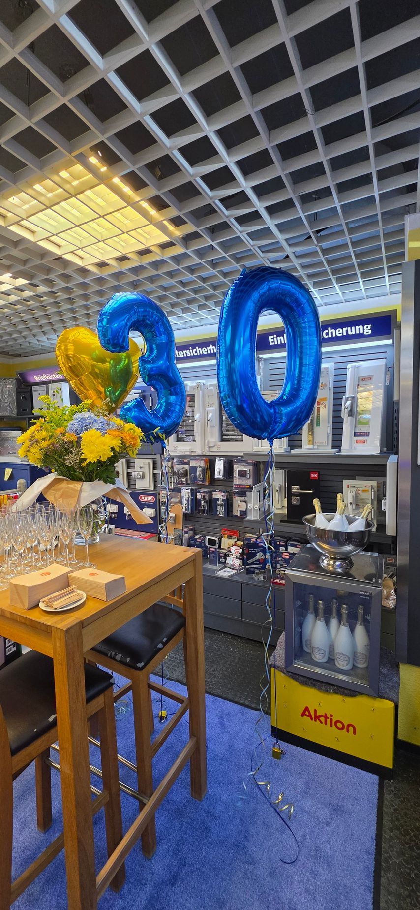 Ein blauer Ballon mit der Zahl „20“ steht in einem Laden mit Kleiderständern und einem Holztisch unter einer Gitterdecke.