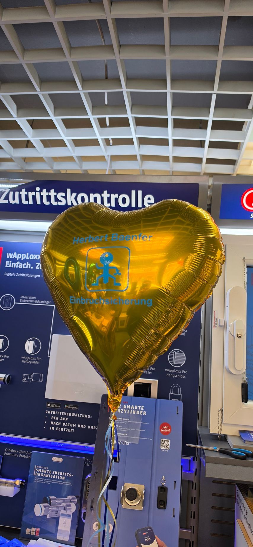 Ein goldener, herzförmiger Ballon im Check-in-Bereich eines Flughafens, im Hintergrund das Schild „Zutrittskontrolle“.