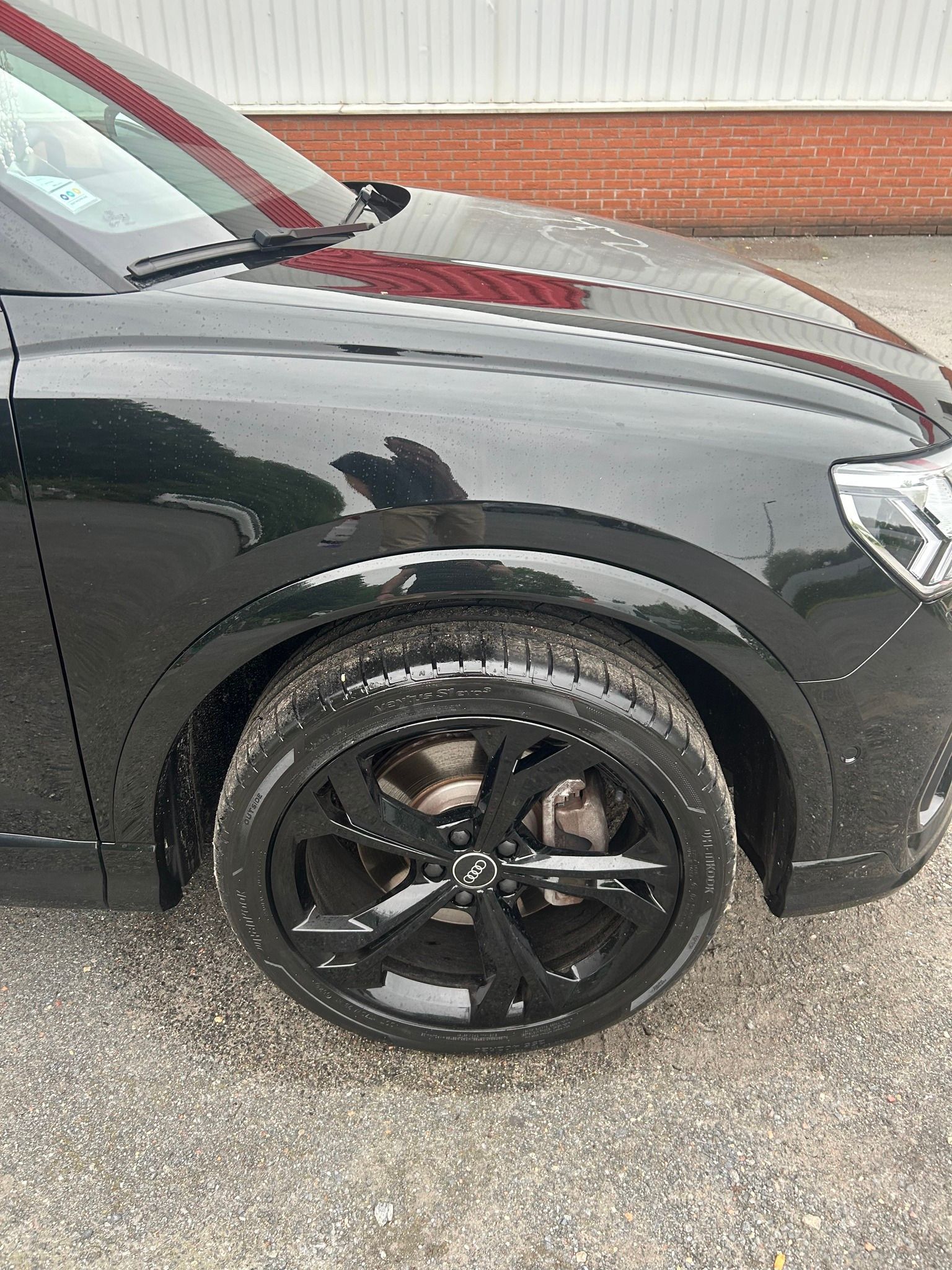 Voiture noire, pneu avant et garde-boue sur gravier. La voiture a des jantes noires et est garée près d'un bâtiment.