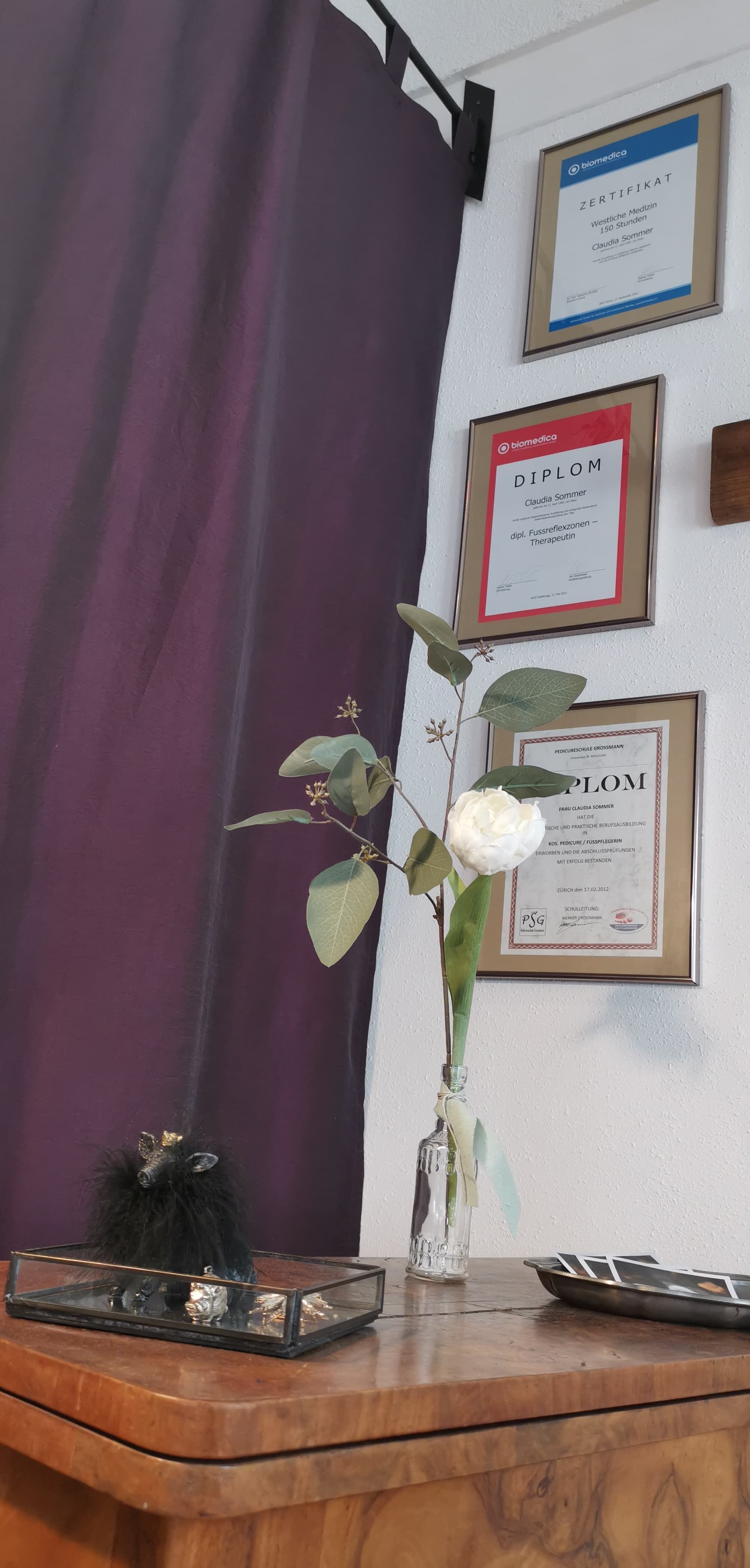 Eine weiße Rose in einer Glasvase steht auf einem Holzschrank mit einem Aschenbecher und gerahmten Dokumenten vor einer weißen Wand.