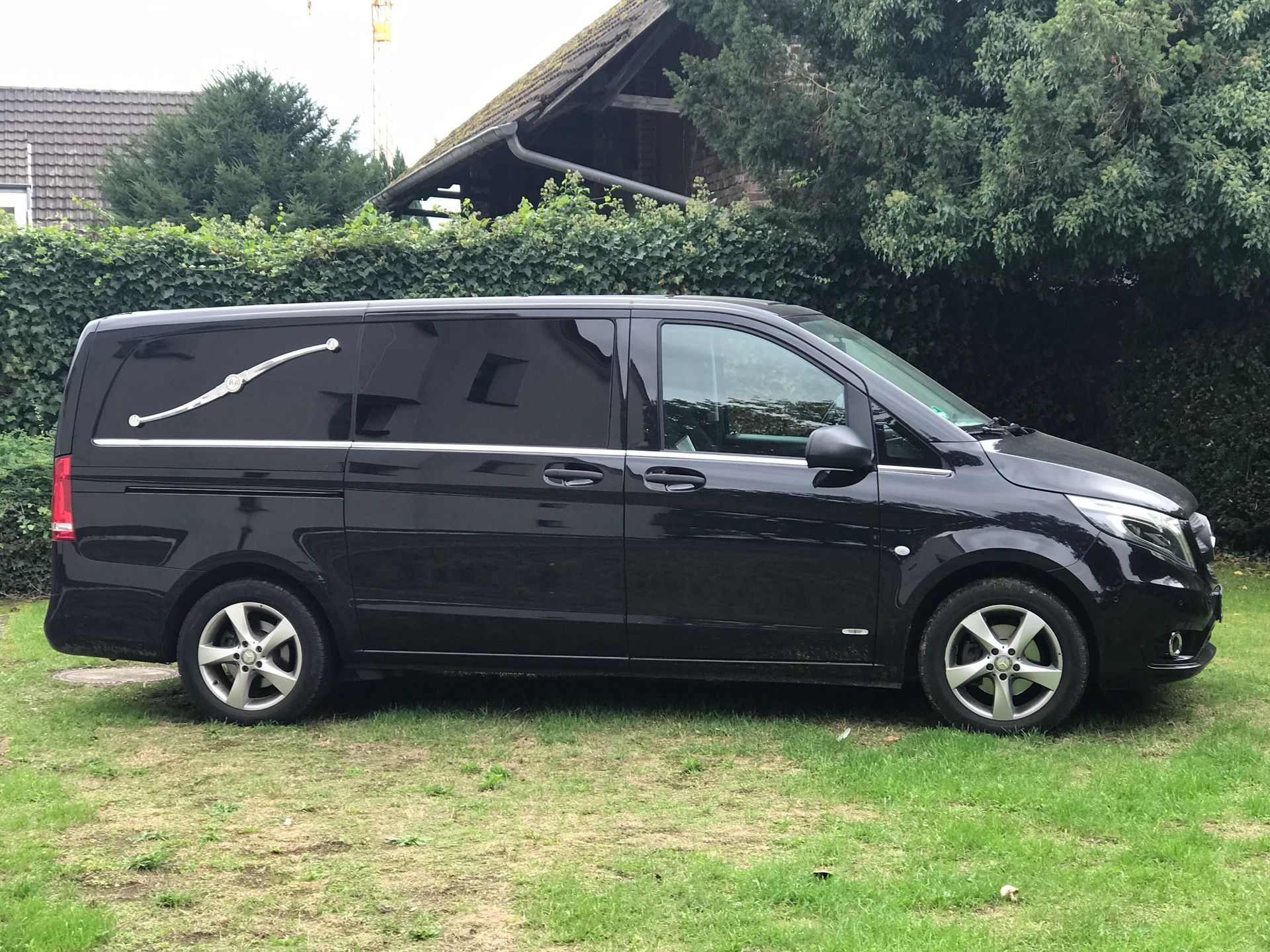 Schwarzer Leichenwagen auf Gras geparkt, mit einem Haus und Bäumen im Hintergrund.