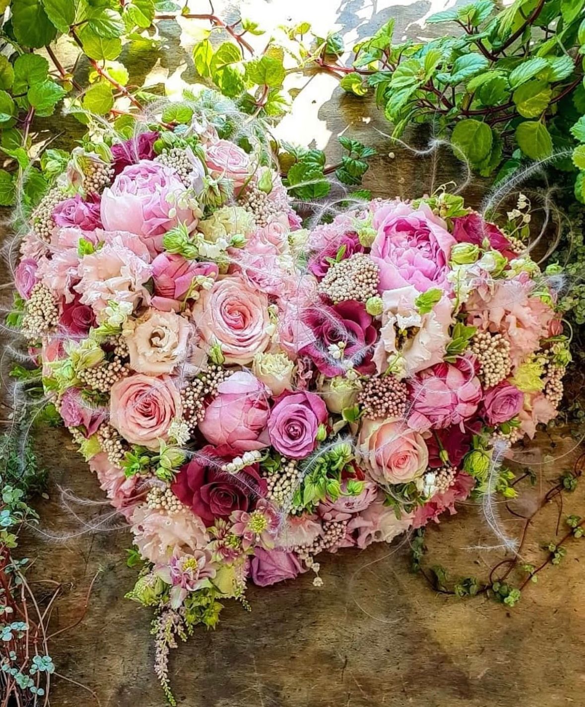 Herzförmiges Blumenarrangement aus rosa, violetten und grünen Blüten auf einem strukturierten braunen Untergrund.
