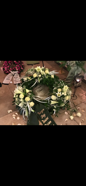Ein Blumenkranz mit weißen Blumen und einem schwarzen Band, wahrscheinlich bei einer Beerdigung.