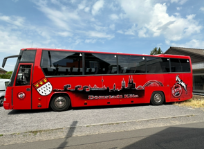 Roter Bus mit FC Köln-Grafiken, im Freien geparkt. Mit Stadtbild und Vereinslogo.