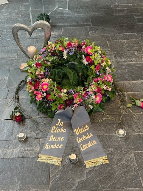 A colorful floral wreath with a ribbon that reads "In Liebe Deine Kinder" and "Ulrich, Carola, Carolin."