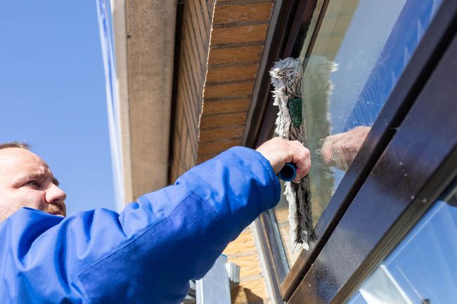Ein Mann in einer blauen Jacke putzt ein Fenster mit einer Bürste.