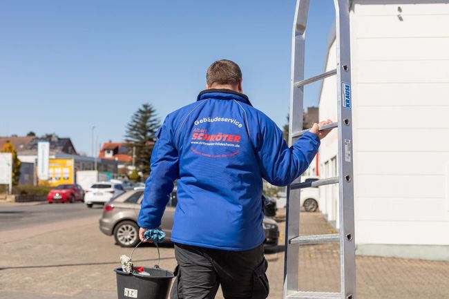 Ein Mann in einer blauen Jacke trägt eine Leiter und einen Eimer.