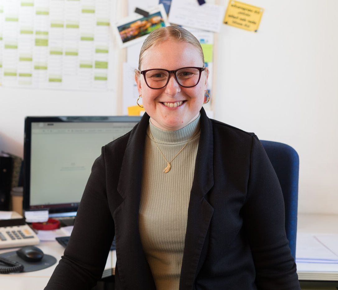 Eine Frau mit Brille sitzt an einem Schreibtisch vor einem Computer.