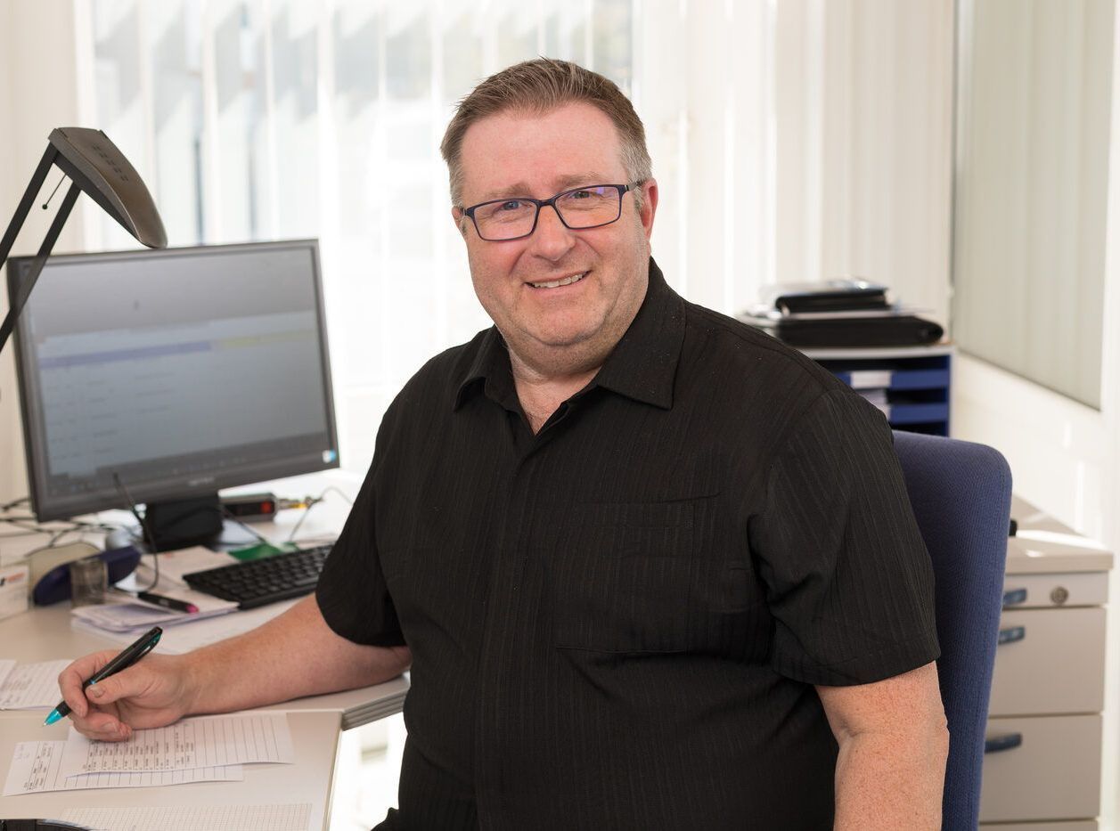 Ein Mann in einem schwarzen Hemd sitzt an einem Schreibtisch vor einem Computer.