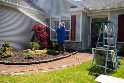 Une personne vêtue d'une chemise bleue nettoie au jet d'eau la façade d'une maison grise.