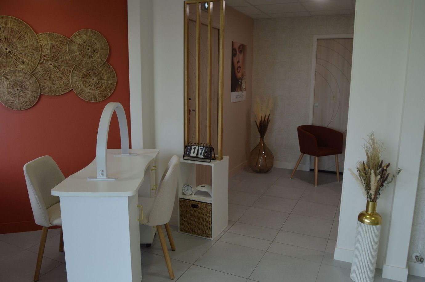 Un espace dédié aux ongles comprenant une table de manucure blanche, deux chaises, un mur en terre cuite et des éléments décoratifs floraux séchés.