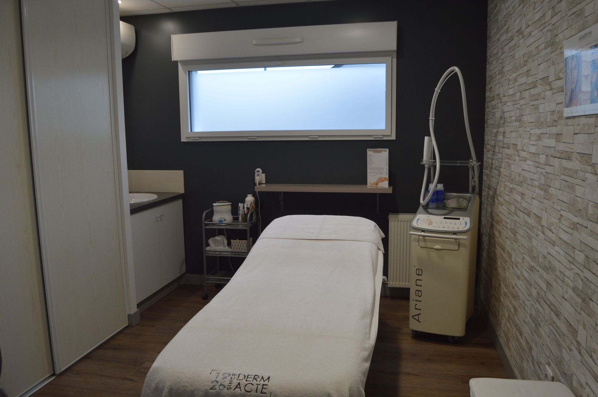 Une salle de soins avec une table de massage blanche, un petit lavabo, un chariot de rangement à roulettes et du matériel de beauté.