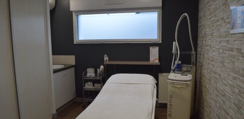 Une salle de soins avec une table de massage blanche, un petit lavabo, un chariot de rangement à roulettes et du matériel de beauté.