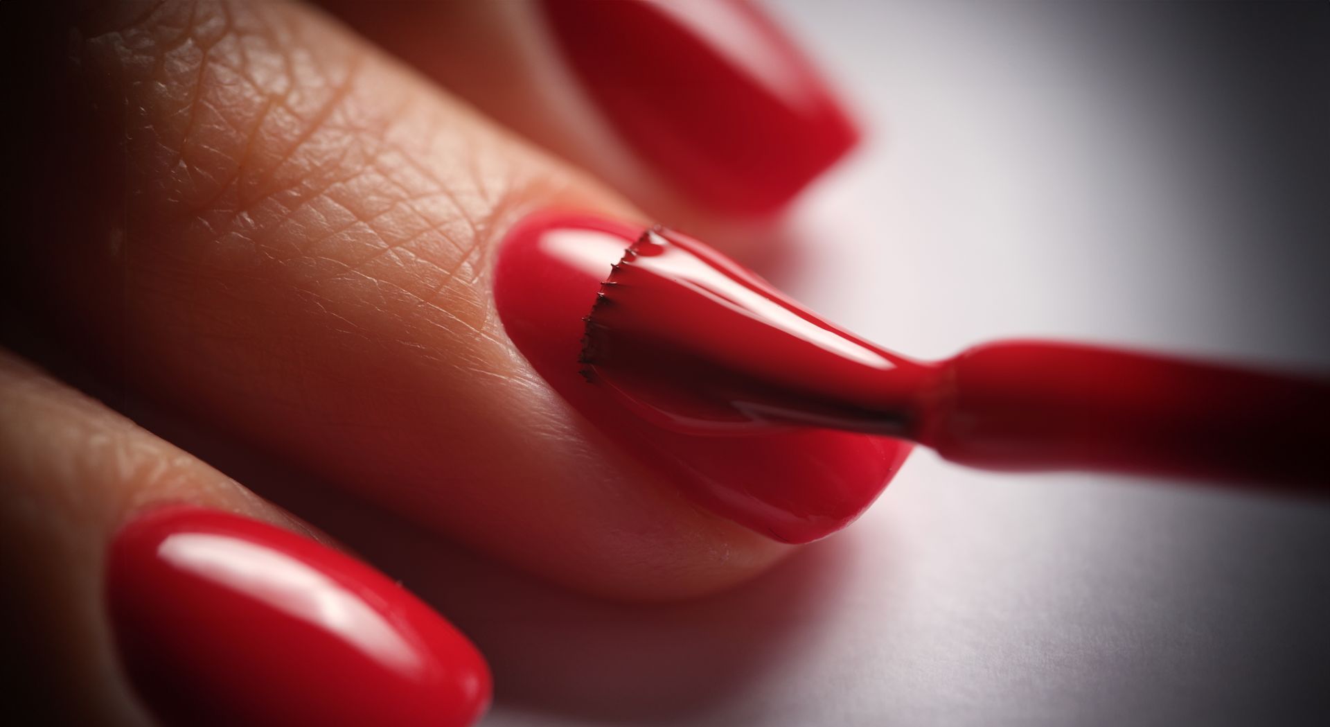 Gros plan sur un pinceau appliquant du vernis à ongles rouge vif sur un ongle, avec l'ongle rouge fini visible en dessous.