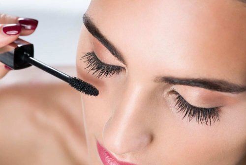 Une main aux ongles vernis d'un rouge foncé utilise une brosse à mascara pour appliquer du maquillage noir sur les cils d'une personne.