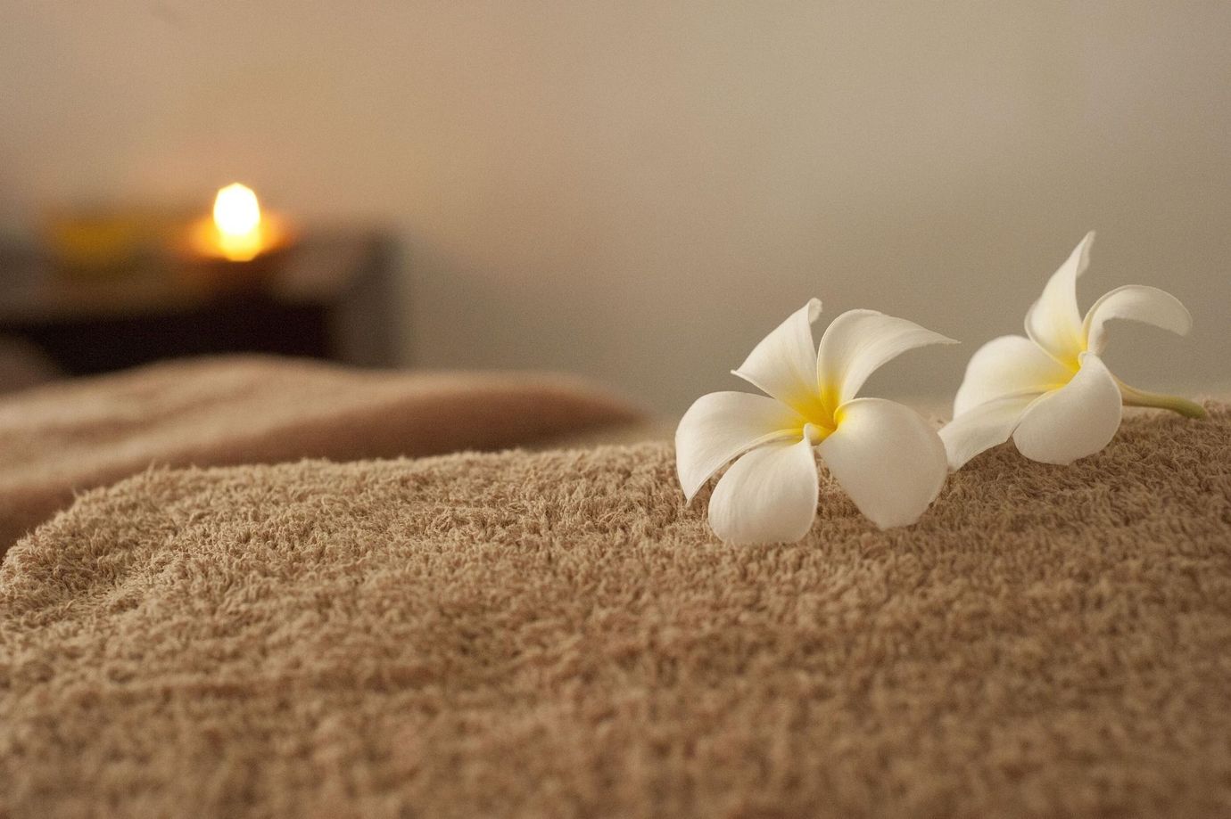 Deux fleurs de frangipanier blanches reposent sur une serviette beige soigneusement pliée, dans une ambiance de spa tamisée, éclairé par une bougie.