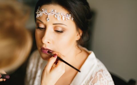 Gros plan sur une personne se faisant maquiller les lèvres au pinceau fin, vêtue d'une robe blanche et d'une coiffe florale.