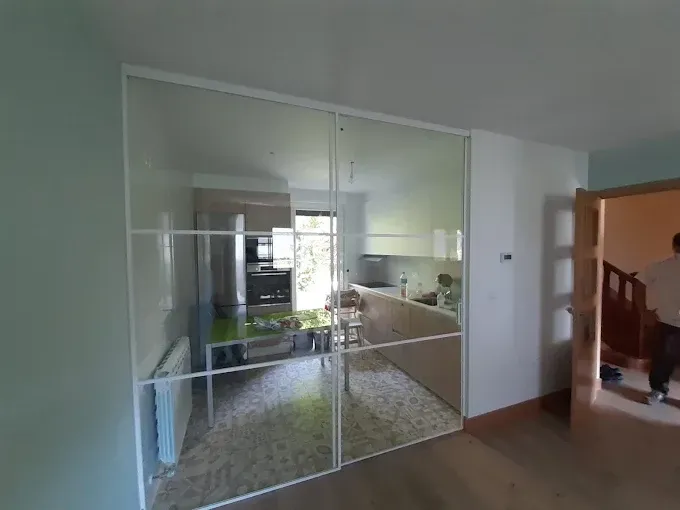 Una mampara corredera minimalista de cristal y marco blanco separa la cocina de la sala de estar, visible desde un pasillo.