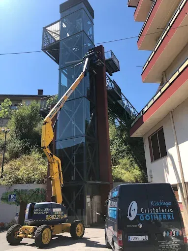 Un ascensor se extiende hacia un hueco de ascensor exterior alto y acristalado, una furgoneta de Cristalería Goiherri.