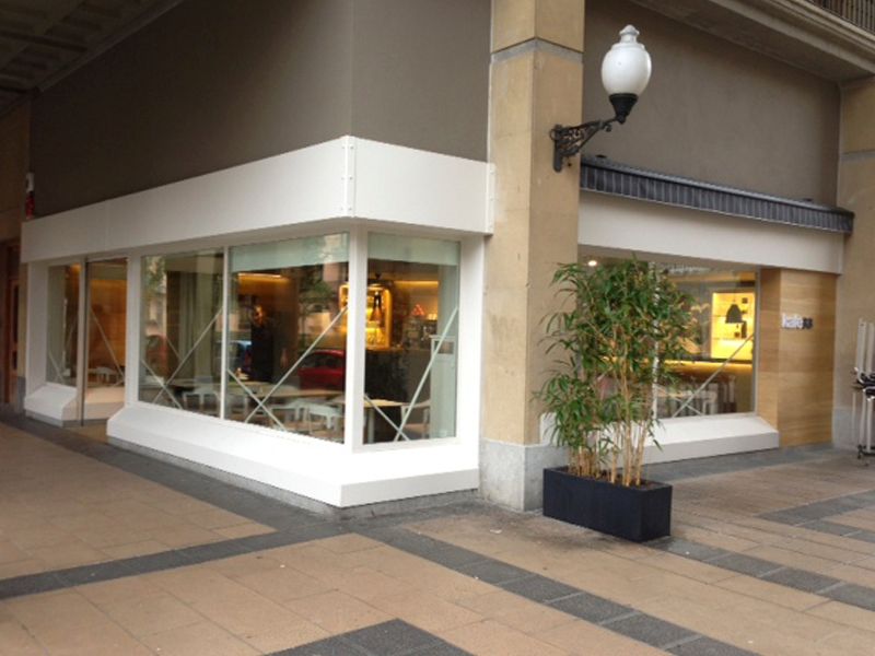 Una tienda de barrio a pie de calle, con marcos de ventanas blancos y una gran planta en maceta cerca de la entrada