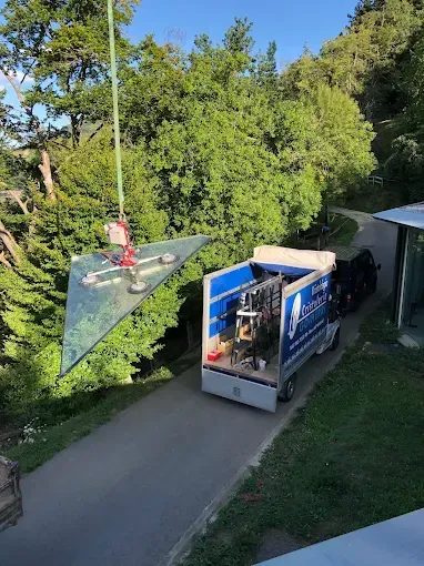 Un panel de vidrio triangular cuelga suspendido de una grúa, que lo iza desde un camión en una carretera pavimentada 