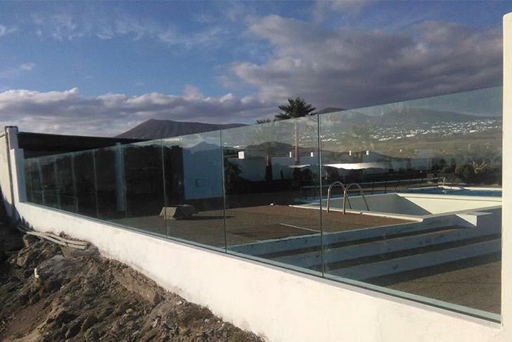 Una valla de cristal rodea una piscina con una casa al fondo.