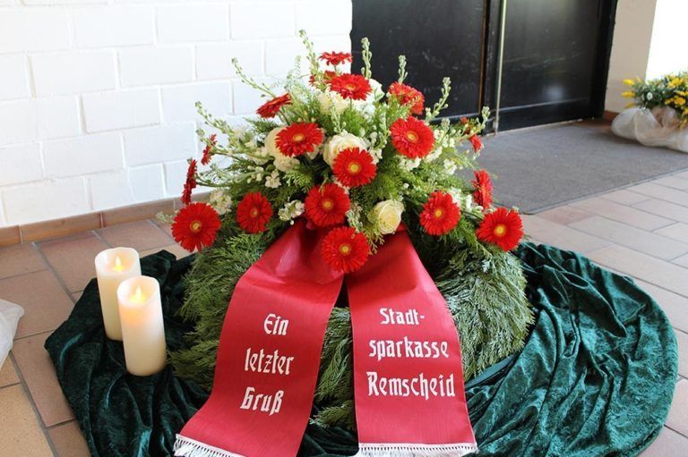 Herzförmiges Blumenarrangement mit roten Rosen und Schleifen bei einer Trauerfeier.