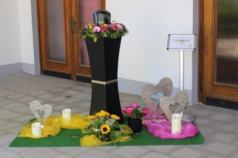 Urnenaufstellung mit Blumen, Kerzen und farbigen Tüchern in einem Innenraum für eine Gedenkfeier.