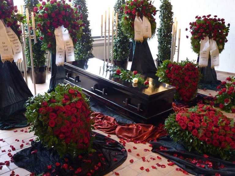 Sarg umgeben von Blumenarrangements und Kränzen im festlich dekorierten Raum.