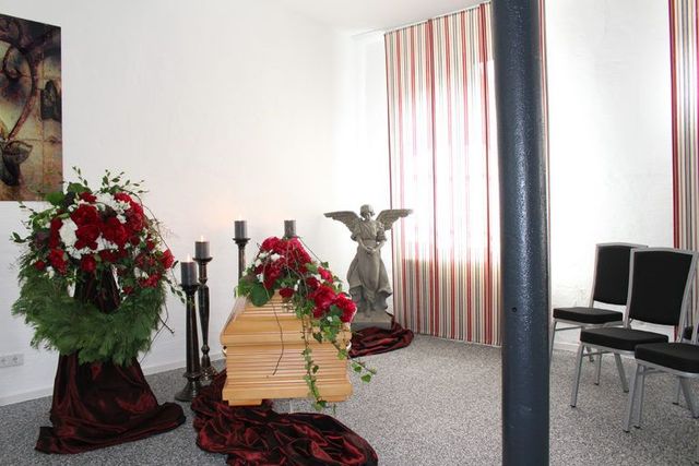 Kleiner Abschiedsraum mit Sarg, Blumenschmuck und Sitzgelegenheiten im Hintergrund.