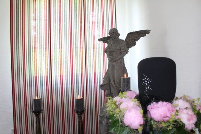 Urne neben Engelsskulptur und Blumen in einem hellen Raum mit gestreiftem Vorhang.