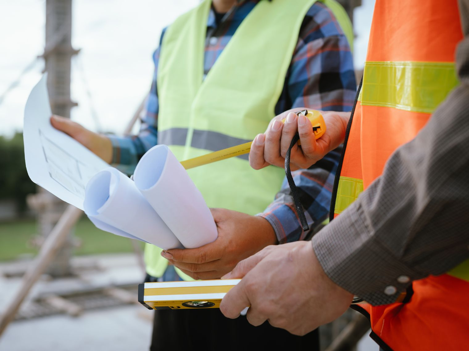 Deux ouvriers du bâtiment examinent des plans, l'un d'eux tenant un mètre ruban, à l'extérieur, vêtus de gilets de sécurité.