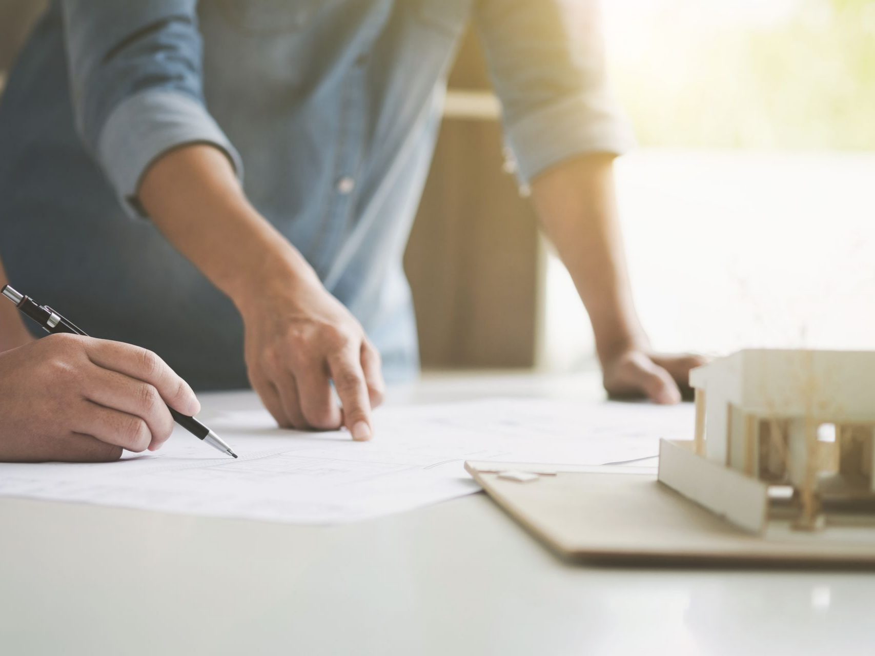 Deux personnes examinent des plans architecturaux, avec une maison témoin à proximité.