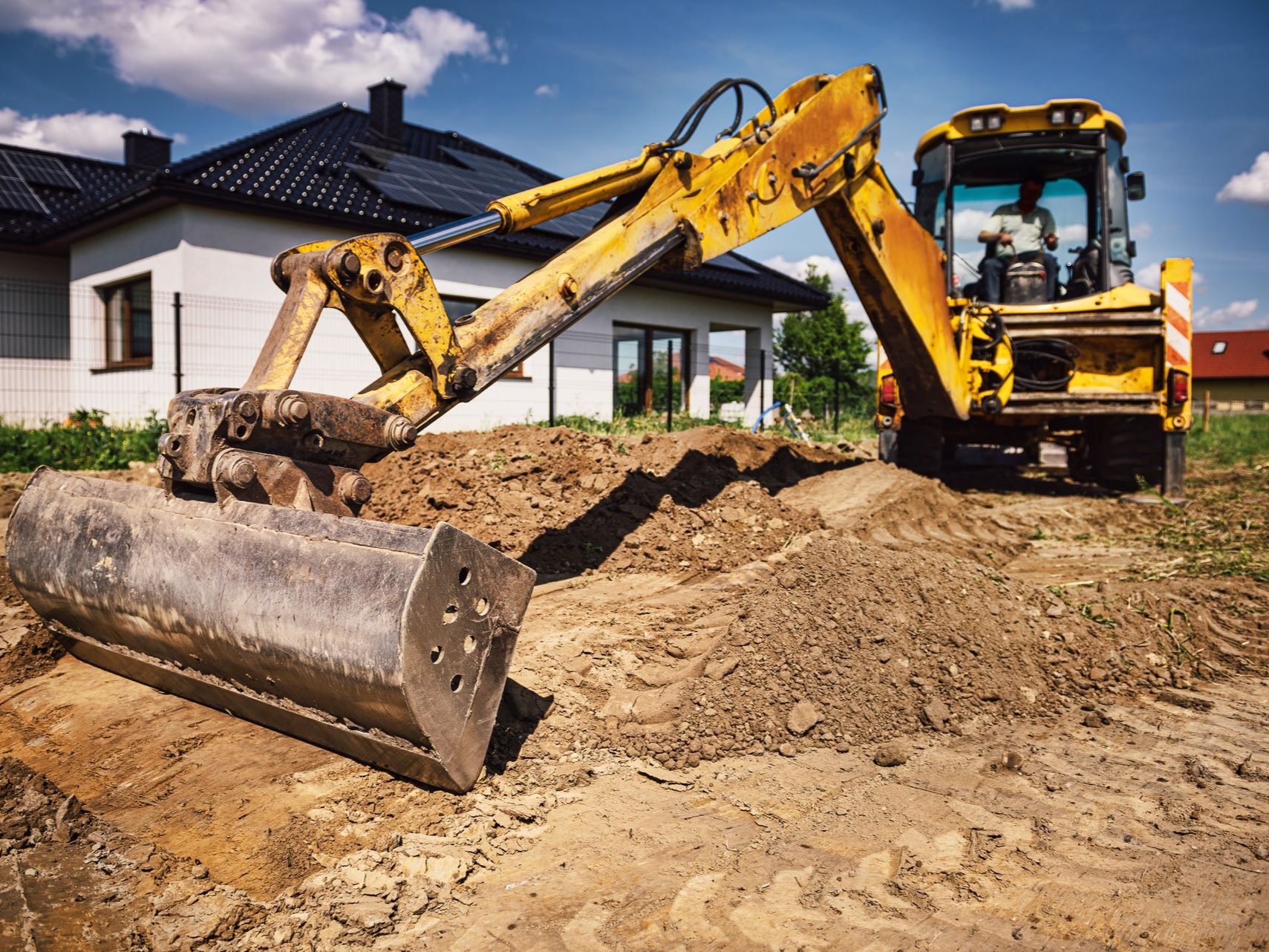 Une pelleteuse jaune creuse une tranchée devant une maison.