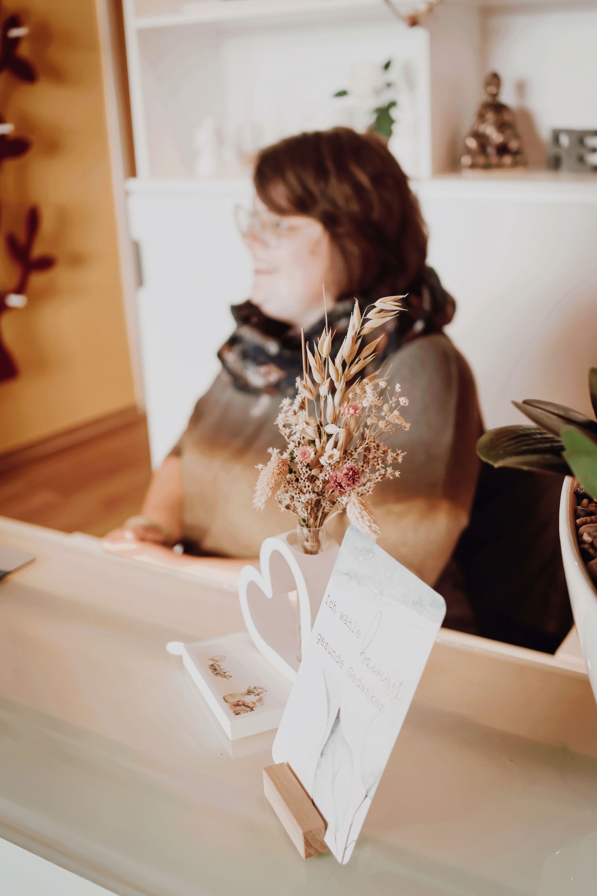 Eine Frau sitzt an einem Schreibtisch vor einer Vase mit getrockneten Blumen.