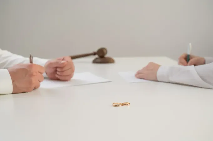 Dos personas firman documentos en una mesa; entre ellas hay anillos de boda. Un mazo reposa al fondo.
