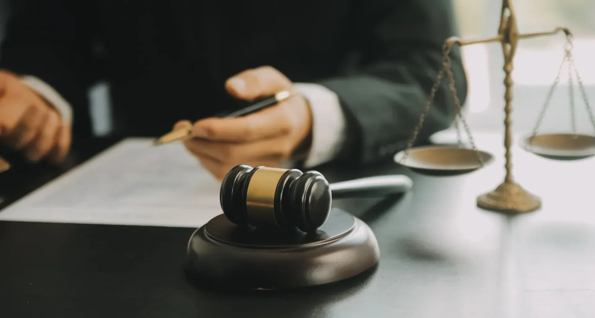 Mazo sobre un atril, balanza de la justicia, persona escribiendo en papel. Oficina legal, colores dorados y oscuros.