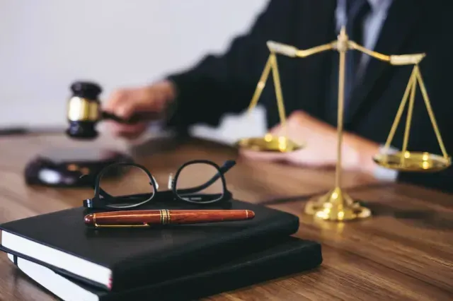 Primer plano: balanza de la justicia, mazo, gafas, bolígrafo, libros sobre el escritorio con una persona al fondo.