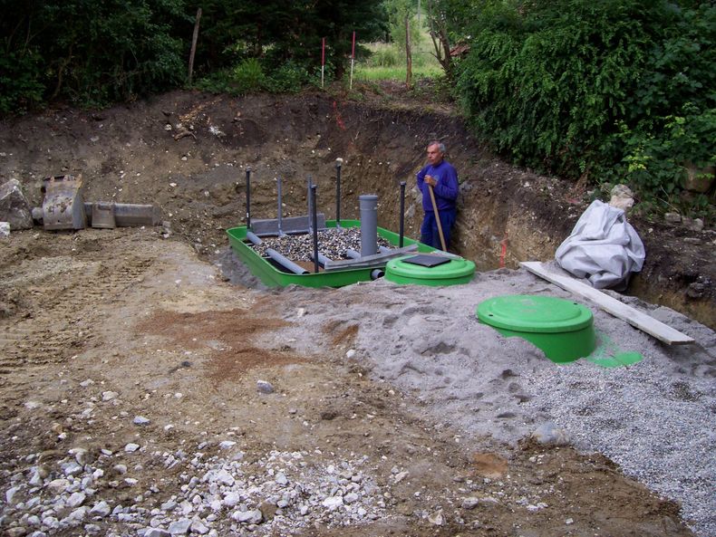 Chantier de construction avec des réservoirs verts et deux ouvriers près d'une fosse creusée dans une zone boisée.
