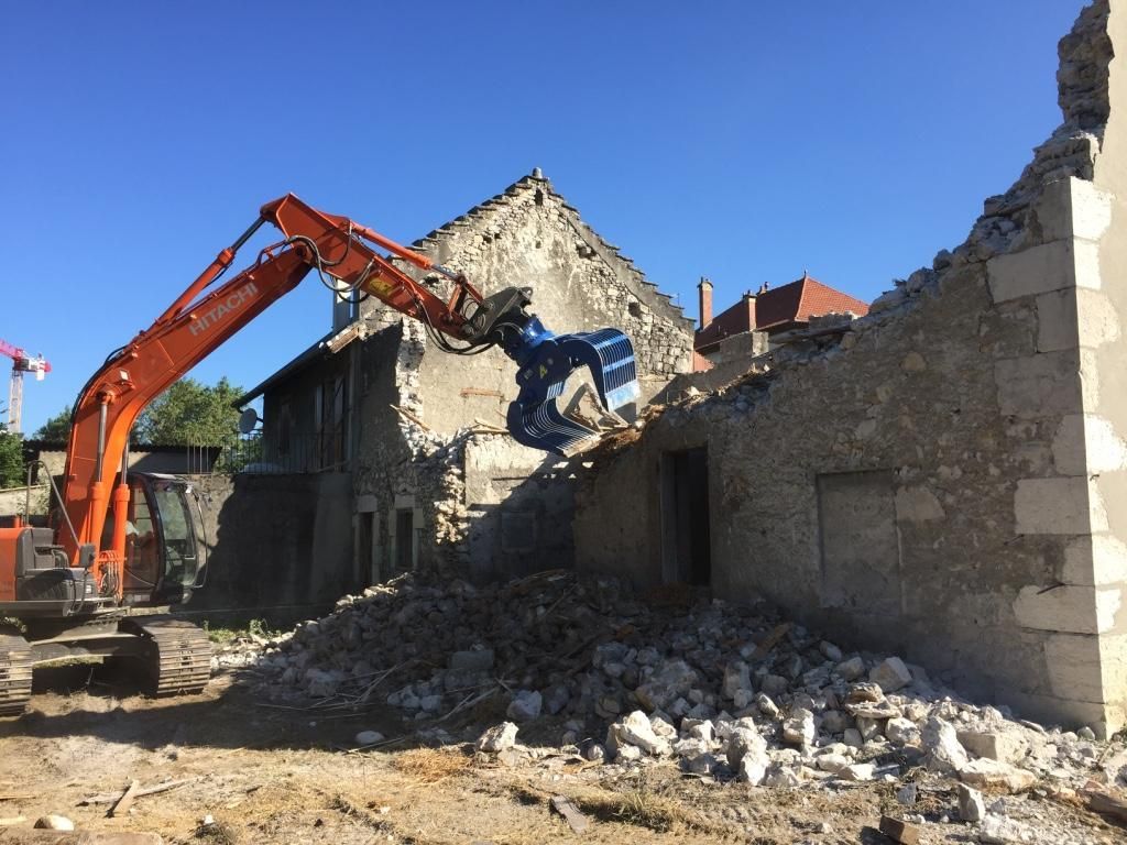 Une pelleteuse orange démolit un bâtiment en pierre sous un ciel bleu clair.