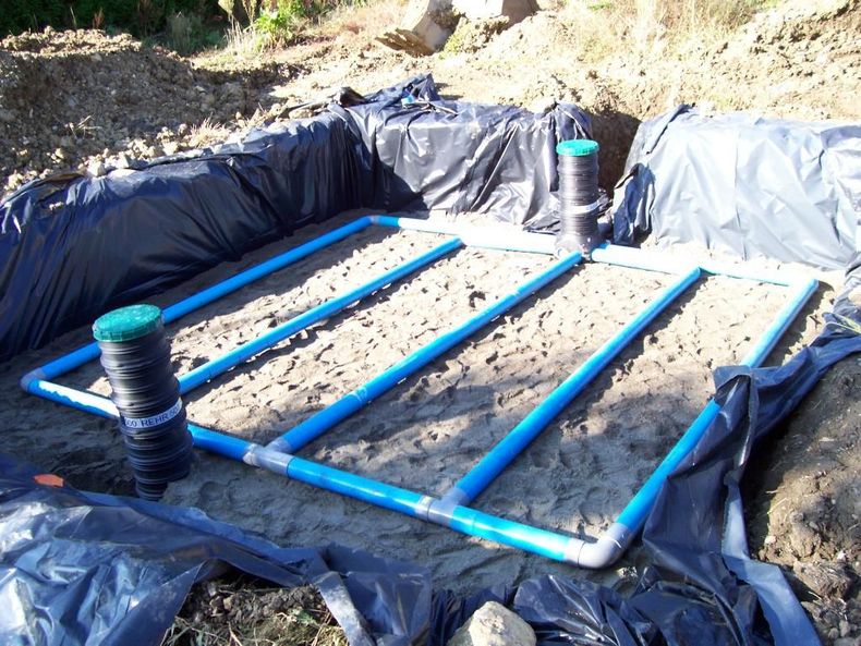 Une petite structure de bassin recouverte d'une bâche, avec des tuyaux en PVC bleus et des supports noirs, installée à l'extérieur sur un sol sablonneux.