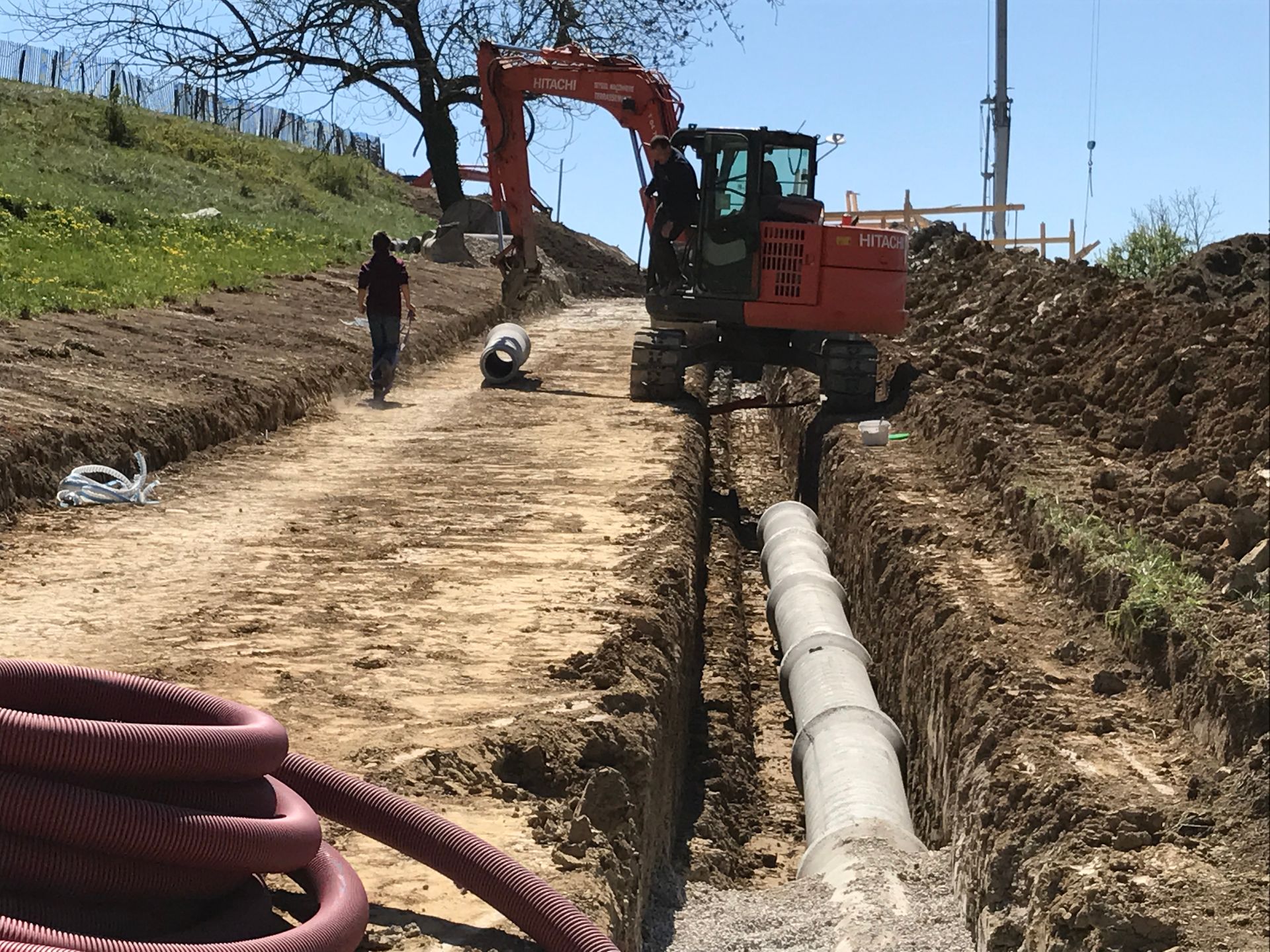 Une excavatrice pose un gros tuyau dans une tranchée de terre sur un chantier de construction, en présence d'ouvriers.