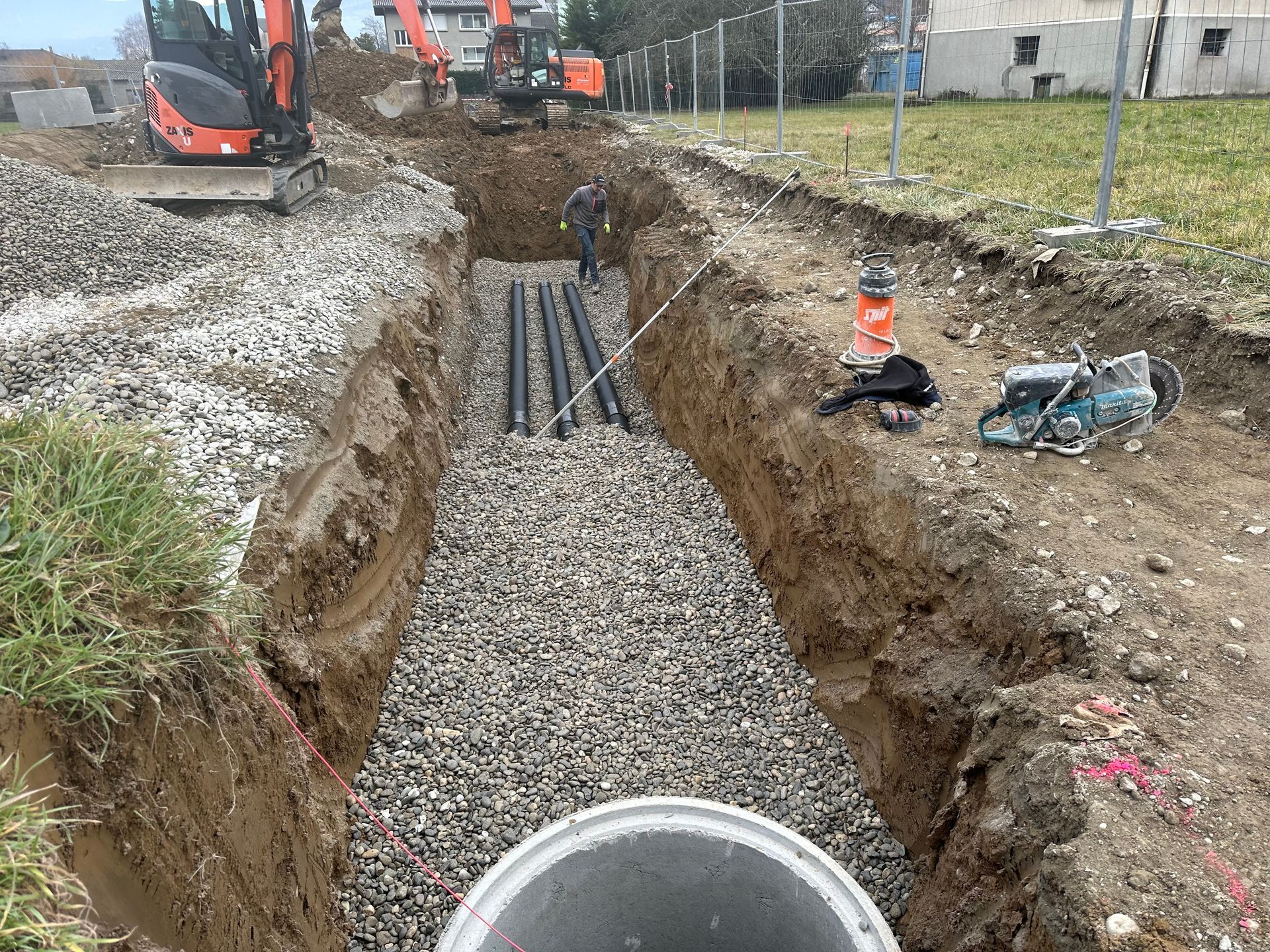 Tranchée de construction avec fond de gravier, tuyaux noirs et excavatrices en arrière-plan.
