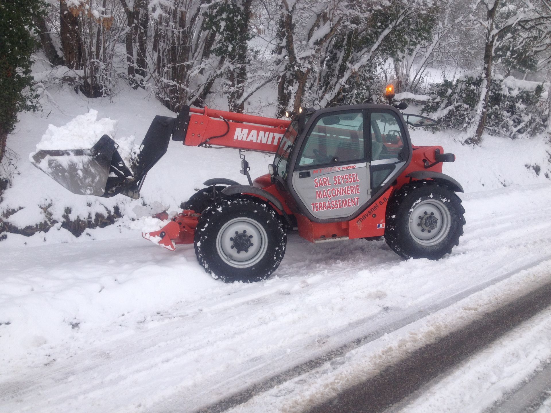 Tracteur rouge avec chargeur frontal déneigeant le bord d'une route enneigée dans une zone boisée.