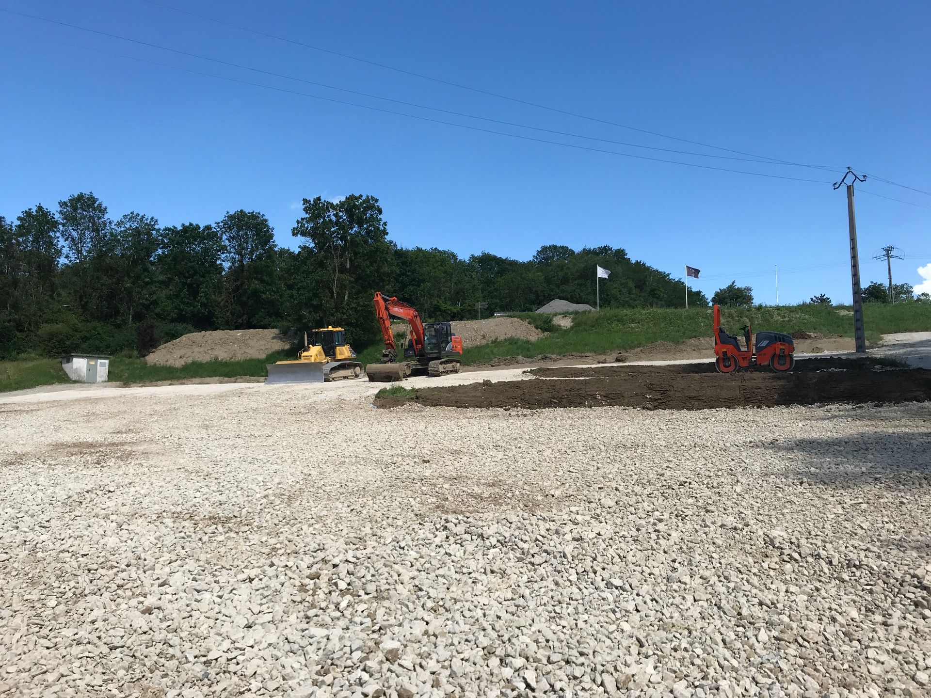 Chantier de construction avec une excavatrice sur un terrain gravillonné sous un ciel bleu, avec des arbres et des poteaux électriques en arrière-plan.