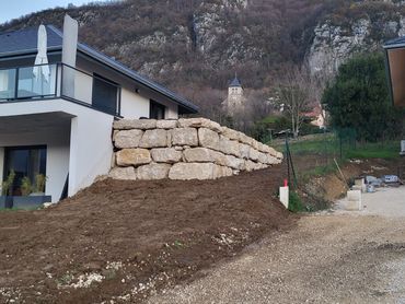 Mur de soutènement en pierres empilées à côté d'une maison blanche sur un talus de terre dans un cadre rural vallonné.